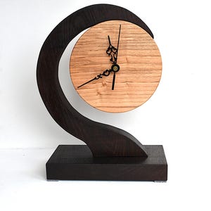 May include: A table clock featuring a circular light-toned wood face with black ornate hands, suspended within a dark-toned curved wooden frame that rises from a rectangular base. The design showcases a contrast between the light and dark wood.