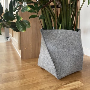 Può includere: Un vaso in feltro grigio con una pianta verde a foglie. Il vaso è appoggiato su un pavimento di legno.