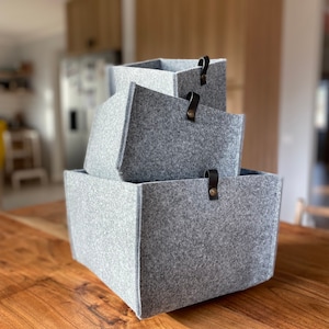 Peut inclure: Trois boîtes de rangement en feutre gris avec des poignées en cuir noir. Les boîtes sont empilées les unes sur les autres sur une surface en bois.