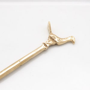 Könnte beinhalten: Ein goldfarbener Metallrührstab mit einem vogelartigen Griff. Der Rührstab liegt auf einem weißen Teller mit goldfarbenem Rand.