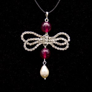 May include: A silver-toned bow pendant necklace with clear crystals. The necklace features two round, deep red beads and a teardrop-shaped pearl. The pendant hangs from a black cord against a black backdrop.