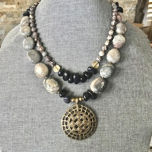 May include: A layered necklace with large grey and tan stone beads, small black beads, and a gold-toned pendant with a spiral design. The necklace is displayed on a grey surface.