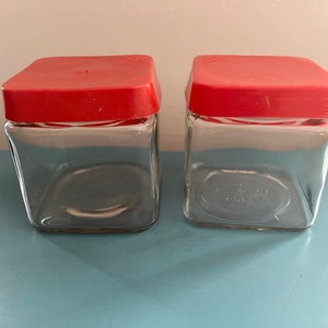May include: Two square glass jars with red lids. The jars are clear glass and the lids are a bright red.