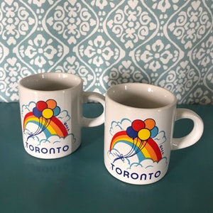May include: Two white ceramic mugs with a handle. Each mug features a rainbow, balloons, and the word "TORONTO" in blue. The design is set against a cloud background. The mugs are on a blue surface.