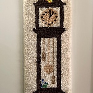 May include: A vintage, handmade wall hanging featuring a brown grandfather clock design. The clock face is tan with black markings, and the pendulum and weights are visible. A small cat is at the base. The background is a textured, off-white material.