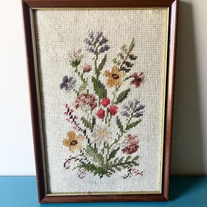 May include: A rectangular cross-stitch embroidery featuring a colourful floral bouquet with yellow, purple, red, and green threads on a cream-coloured fabric background, encased in a dark brown wooden frame with a thin gold-toned inner border.