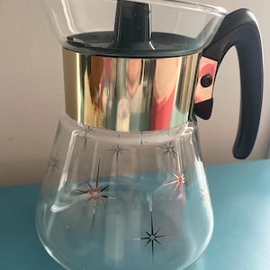 May include: Vintage glass coffee carafe with a clear spout and a black handle. The carafe features a gold band and a dark green lid. The glass is decorated with starburst designs in gold and black. A retro kitchenware item.