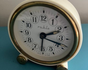 May include: A white alarm clock with gold accents and black numbers. The clock face reads "ruhla" and "MADE IN GDR".