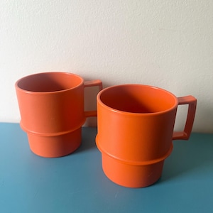 May include: Two bright orange mugs with a textured finish and a distinct horizontal ridge around the middle. Each mug features a square-shaped handle and a cylindrical body designed for stacking, showcasing a uniform vibrant color throughout.