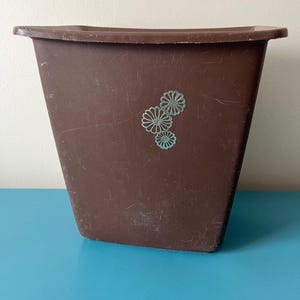 May include: A brown plastic wastebasket with a slightly flared shape. Three turquoise flower designs are on the front. The wastebasket has a rectangular top and is set against a blue background.