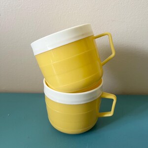 May include: Two stacked mugs featuring a bright yellow body with horizontal embossed lines and a contrasting solid white rim. Each mug includes a matching yellow angular handle attached to the side, creating a uniform appearance for the set.