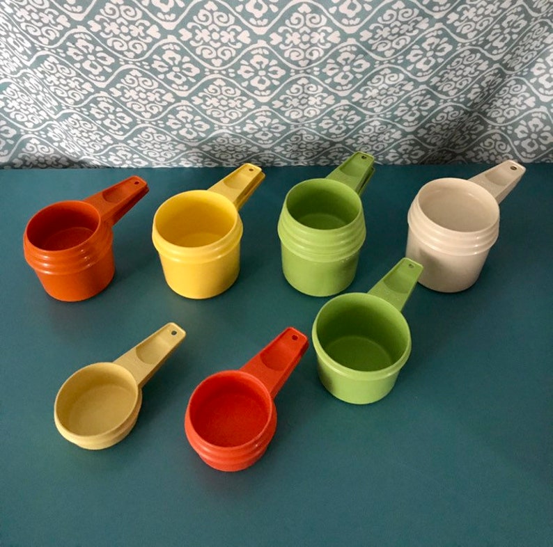 May include: Set of seven vintage plastic measuring cups in various colours, including orange, yellow, green, and cream. The cups are nested together in a stack.