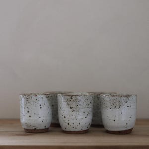 May include: A set of five ceramic cups with a white glaze and dark brown speckles. Each cup features a cylindrical shape with a slightly tapered base and an exposed, unglazed brown clay bottom, arranged in a cluster on a flat surface.