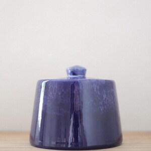 Puede incluir: Un pequeño tarro de cerámica azul oscuro con tapa. El tarro tiene forma trapezoidal redondeada y acabado brillante. La tapa tiene un pequeño pomo en la parte superior. El tarro está sobre una superficie de madera.