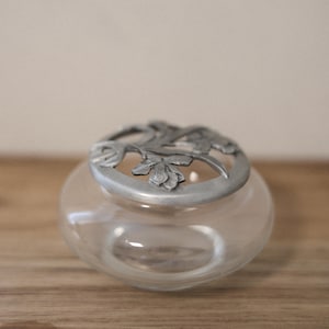 Peut inclure: Un pot en verre transparent avec un couvercle décoratif argenté orné d'un motif floral. Le pot est rond et posé sur une surface en bois. Le couvercle présente un motif ajouré détaillé de feuilles et de fleurs.