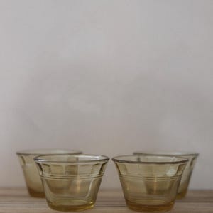 May include: Four amber-colored, vintage-style drinking glasses. Each glass has a slightly flared shape with horizontal bands around the middle. The glasses are arranged on a light-colored wooden surface.