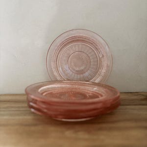 Può includere: Una pila di quattro piatti rotondi in vetro rosa traslucido con un motivo testurizzato a raggiera che si irradia dal centro verso il bordo. I piatti presentano un bordo esterno liscio e rialzato e un delicato design in vetro pressato.