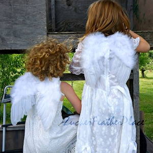 May include: Two children wearing white dresses with white feather angel wings. The wings are attached to the back of the dresses. The children are standing in front of a wooden fence.