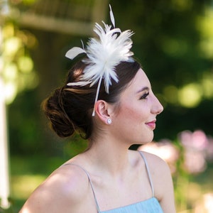 May include: A woman with a white feather headband in her hair, wearing a light blue sleeveless dress. She is looking to the right and smiling.