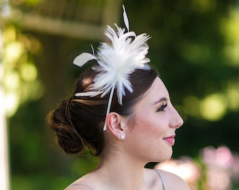 Könnte beinhalten: Ein weißer Feder-Fascinator, bestehend aus einem Bündel zarter, aufrecht stehender Federn, die an einem dünnen, passenden weißen Haarreif befestigt sind. Das Accessoire sitzt seitlich am Kopf und zeigt eine leichte, luftige Textur und ein schlichtes Design.
