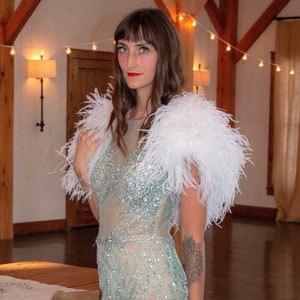 May include: A woman wearing a light blue sequined gown with a white feather shawl. She is standing in a large room with wooden beams and string lights.