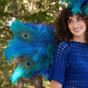 May include: A woman with curly brown hair wears a blue crocheted top and a large headdress made of blue and green feathers. The feathers are arranged in a fan shape and resemble a peacock's tail.