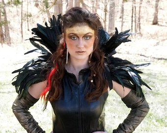 May include: A woman wearing a black leather jacket with a large black feather collar and matching feather shoulder pads. She is wearing black gloves and has gold makeup on her forehead.
