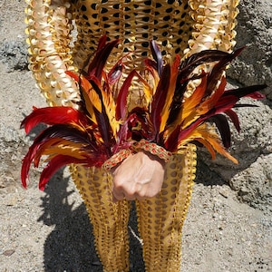 May include: A pair of wrist cuffs featuring vibrant red, orange, and dark brown feathers attached to a gold-toned band with red decorative accents. The cuffs are worn over a gold-coloured, laser-cut, long-sleeved garment with a repeating circular pattern.