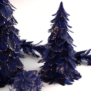Op de afbeelding: Marineblauwe kerstbomen van veren met gouden glitteraccenten. De bomen zijn in verschillende maten en vormen, met een kleine, ronde veerdecoratie op de voorgrond. Op een witte achtergrond.
