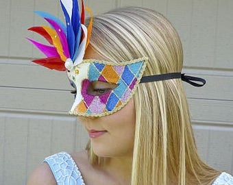 Peut inclure: Un masque de carnaval coloré avec un motif en losange et un panache de plumes. Le masque est blanc avec des accents roses, bleus et oranges.