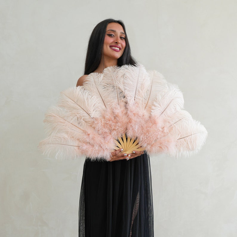 Puede incluir: Una mujer sostiene un gran abanico de plumas rosa y blanco frente a ella. El abanico est&aacute; hecho de plumas suaves y esponjosas y tiene un mango de madera. La mujer lleva un vestido negro.