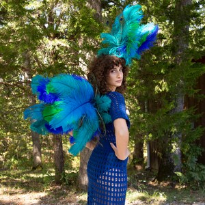 May include: A person wearing a blue crochet dress with a large peacock feather headdress. The headdress is made of blue, green, and teal feathers. The person is standing in a forest setting.