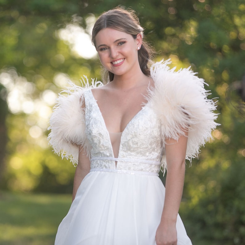 May include: A woman wearing a white dress with a plunging neckline and feather detailing on the shoulders. The dress has a fitted bodice and a flowing skirt.