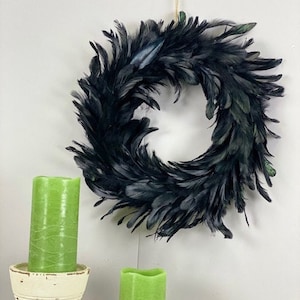 Peut inclure: Une couronne circulaire compos&eacute;e de plumes noires superpos&eacute;es &agrave; l'aspect textur&eacute;. Sous la couronne se trouvent deux bougies cylindriques vert citron, l'une plac&eacute;e dans un bougeoir cr&egrave;me et l'autre pos&eacute;e directement sur la surface.