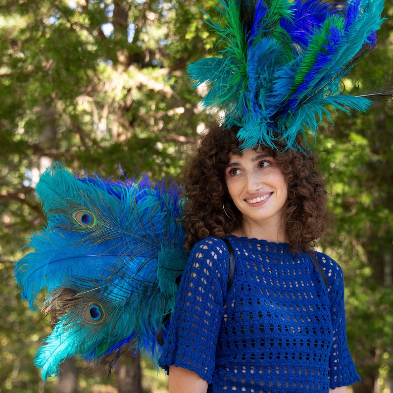 May include: A woman with curly brown hair wears a blue crocheted top and a large headdress made of blue and green feathers. The feathers are arranged in a fan shape and resemble a peacock's tail.