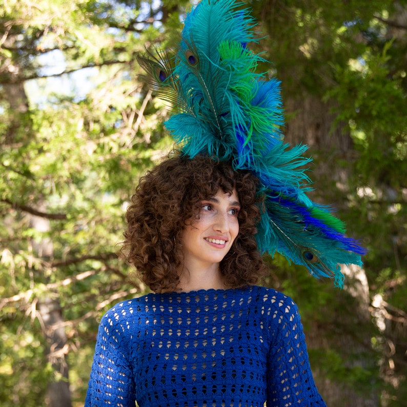 May include: A woman with curly brown hair wearing a blue crocheted top and a large peacock feather headdress.