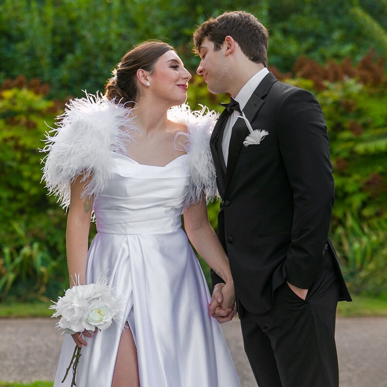 May include: A couple in formal attire, the woman wearing a white wedding dress with a feathered shawl and a bouquet of white flowers, the man wearing a black tuxedo, looking at each other.