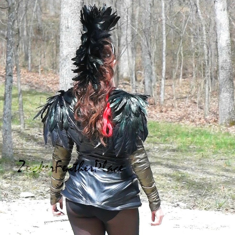 Puede incluir: Una mujer lleva una chaqueta de cuero negra con hombreras de plumas negras y un tocado de plumas negras. La chaqueta tiene detalles dorados en las mangas. La mujer est&aacute; de pie en una zona boscosa.