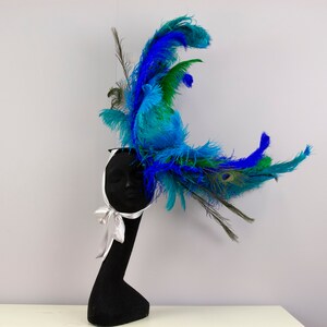 May include: A blue and green feather headpiece with a white satin ribbon. The headpiece is on a black mannequin head.