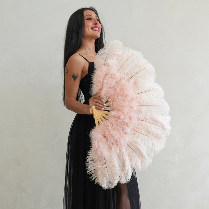 Puede incluir: Una mujer con un vestido negro sostiene un gran abanico de plumas rosa y blanco. El abanico est&aacute; hecho de plumas suaves y tiene un mango de madera.