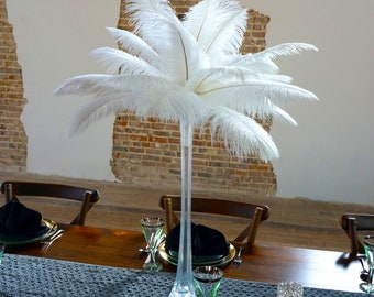 May include: A tall clear glass vase filled with white ostrich feathers, set on a wooden table with a gray table runner and place settings.