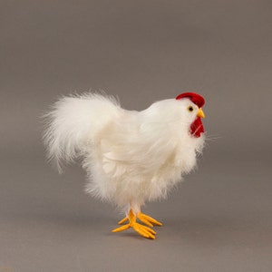 May include: A decorative white chicken figurine with fluffy feathers. It has a red comb and wattle, yellow beak and feet. The chicken is standing on a gray surface.
