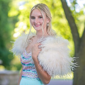 May include: A young person wearing a light blue dress with a white feather boa. The boa is draped over the person's shoulders and is held in place by one hand. The person is smiling and looking at the camera.