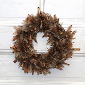 May include: A round wreath made of brown and tan feathers, hanging against a white paneled door. The feathers are arranged densely, creating a full, textured appearance. The wreath is suspended by a thin, natural-colored string.