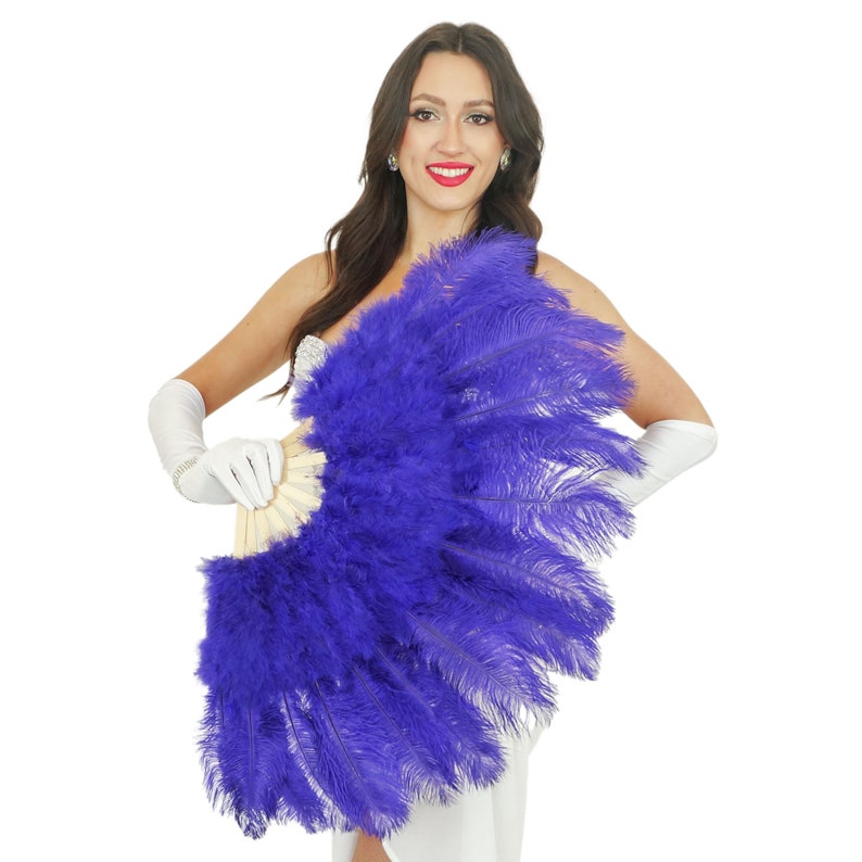 May include: A purple ostrich feather fan with a wooden handle. The fan is held by a person wearing white gloves and a white dress.