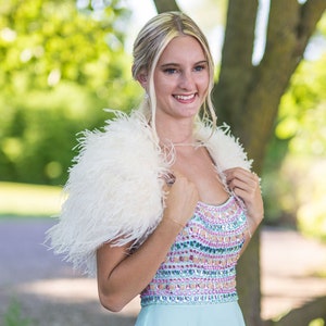 May include: A woman wearing a white feather boa over a light blue dress with a colorful beaded bodice. The dress has a scoop neckline and a fitted bodice.