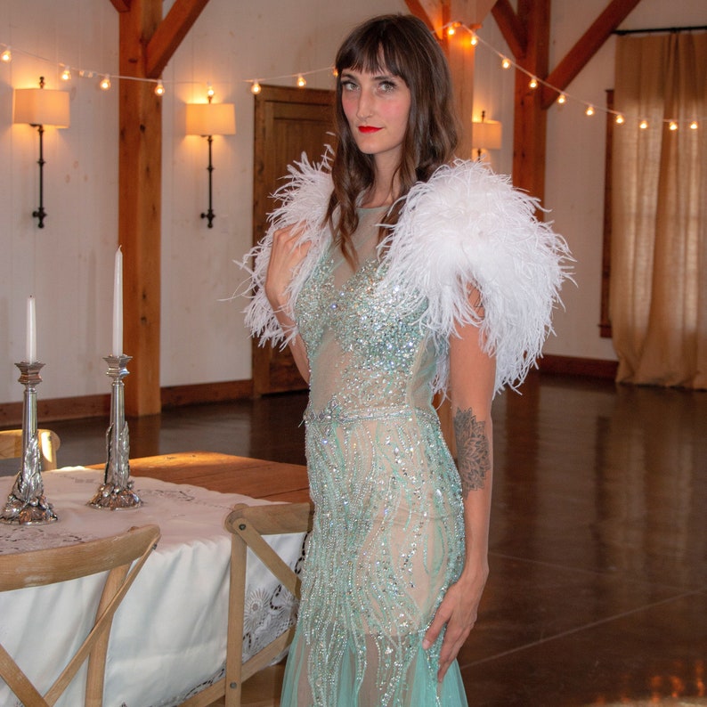 May include: A woman wearing a light blue sequined gown with a white feather shawl. She is standing in a large room with wooden beams and string lights.