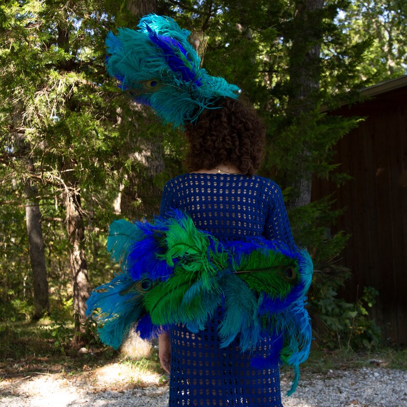 May include: A person wearing a blue crocheted dress with a large peacock feather headdress. The feathers are blue, green, and teal.