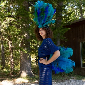 May include: A woman wearing a blue crochet dress with a large blue and green feather headdress. She is standing in a forest setting.