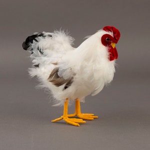 May include: A decorative white rooster figurine with fluffy feathers, a red comb and wattle, and yellow feet. The rooster has black tail feathers and brown accents on its wings. The figurine is standing on a gray surface.